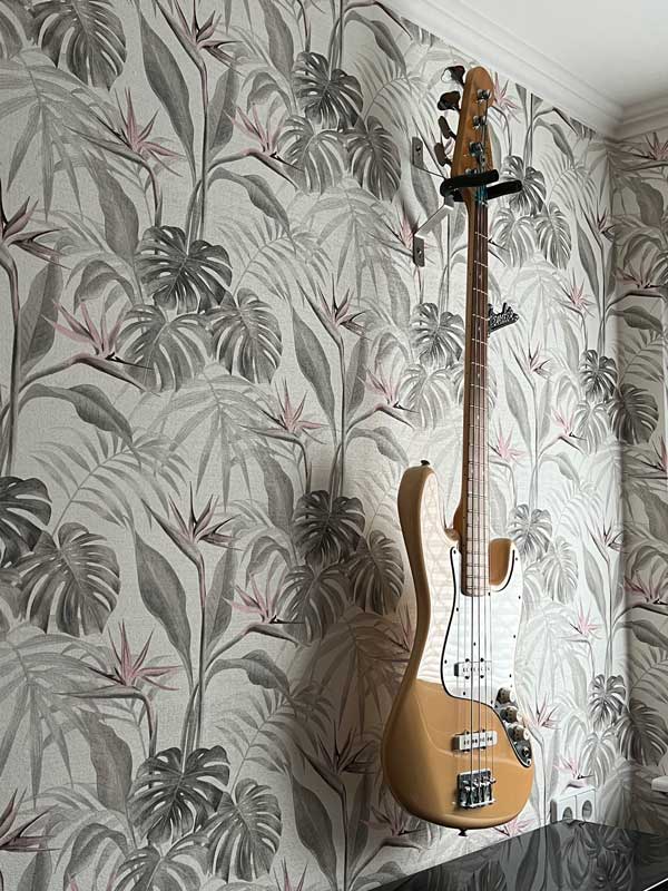 Beige E-Bass-Gitarre, die an einer Wand mit tropisch gemusterter Tapete in gedeckten Farben hängt, kombiniert mit einer schlichten weißen Zierleiste an der Decke.